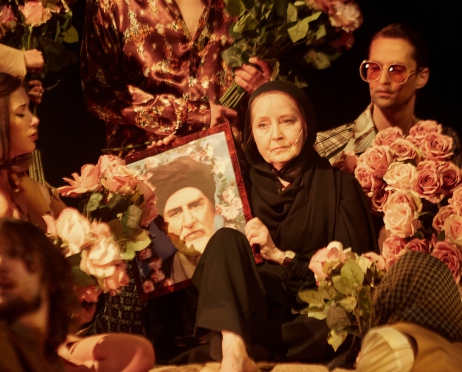 Na zdjęciu kilka osób siedzi blisko siebie. Ubrani są w stylistyce lat siedemdziesiątych. Każdy z nich ma w dłoniach bukiet róż. W centrum siedzi starsza kobieta w czarnej, muzułmańskiej szacie, zakrywającej całe ciało bez twarzy. W dłoniach trzyma fotografię przywódcy.